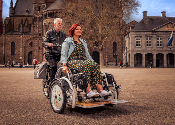 De rolstoelfiets van de Zonnebloem. Een man zit achter het stuur en een vrouw in haar rolstoel voorop de fiets. Ze fietsen door het centrum van Maastricht. 