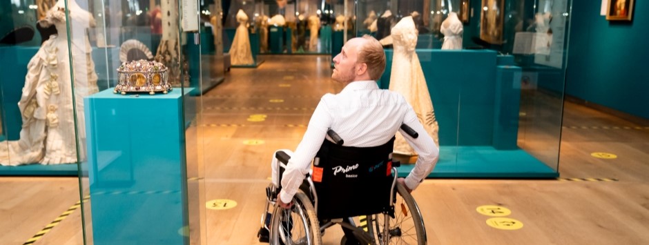Een deelnemer van de belevingsworkshop rolt in een rolstoel door een museum en kijkt naar een vitrine met een kroon.