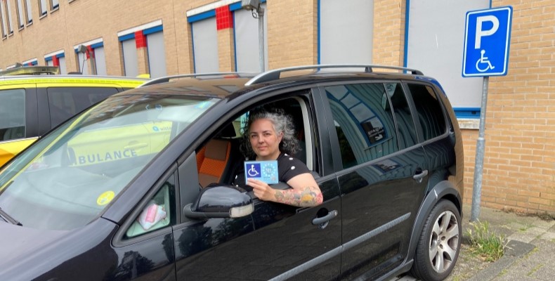 Max in haar auto op invalidenparkeerplaats met gehandiaptenparkeerkaart in haar hand 