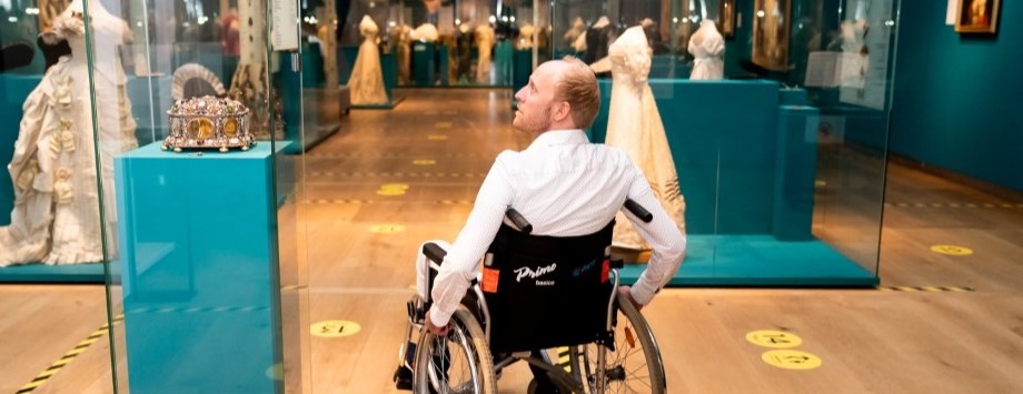 Een deelnemer van de belevingsworkshop rolt in een rolstoel door een museum en kijkt naar een vitrine met een kroon.