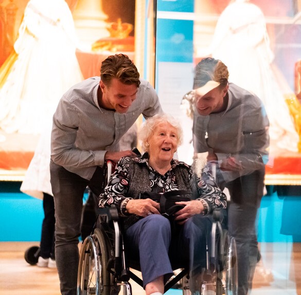 Mannelijke vrijwilliger duwt een rolstoel van een oudere vrouwelijke deelnemer tijdens Het Andere Bedrijfsuitje in een museum