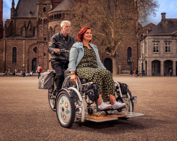 De rolstoelfiets van de Zonnebloem. Een man zit achter het stuur en een vrouw in haar rolstoel voorop de fiets. Ze fietsen door het centrum van Maastricht. 