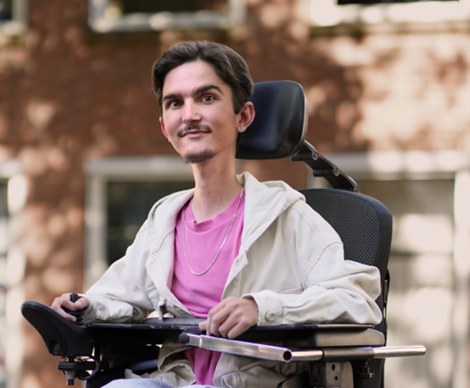 Endri buiten in zijn elektrische rolstoel, glimlachend naar de camera.